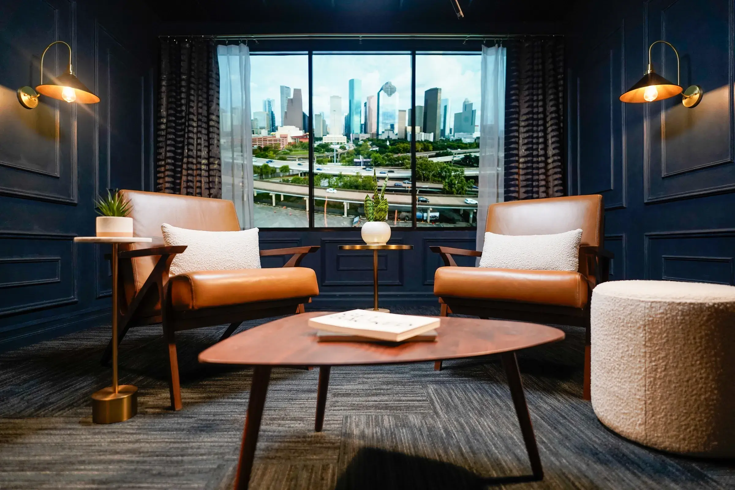 The interview set at rest — two leather chairs and a low table under brass sconces, framed by sheer curtains and heavy drapes at the window