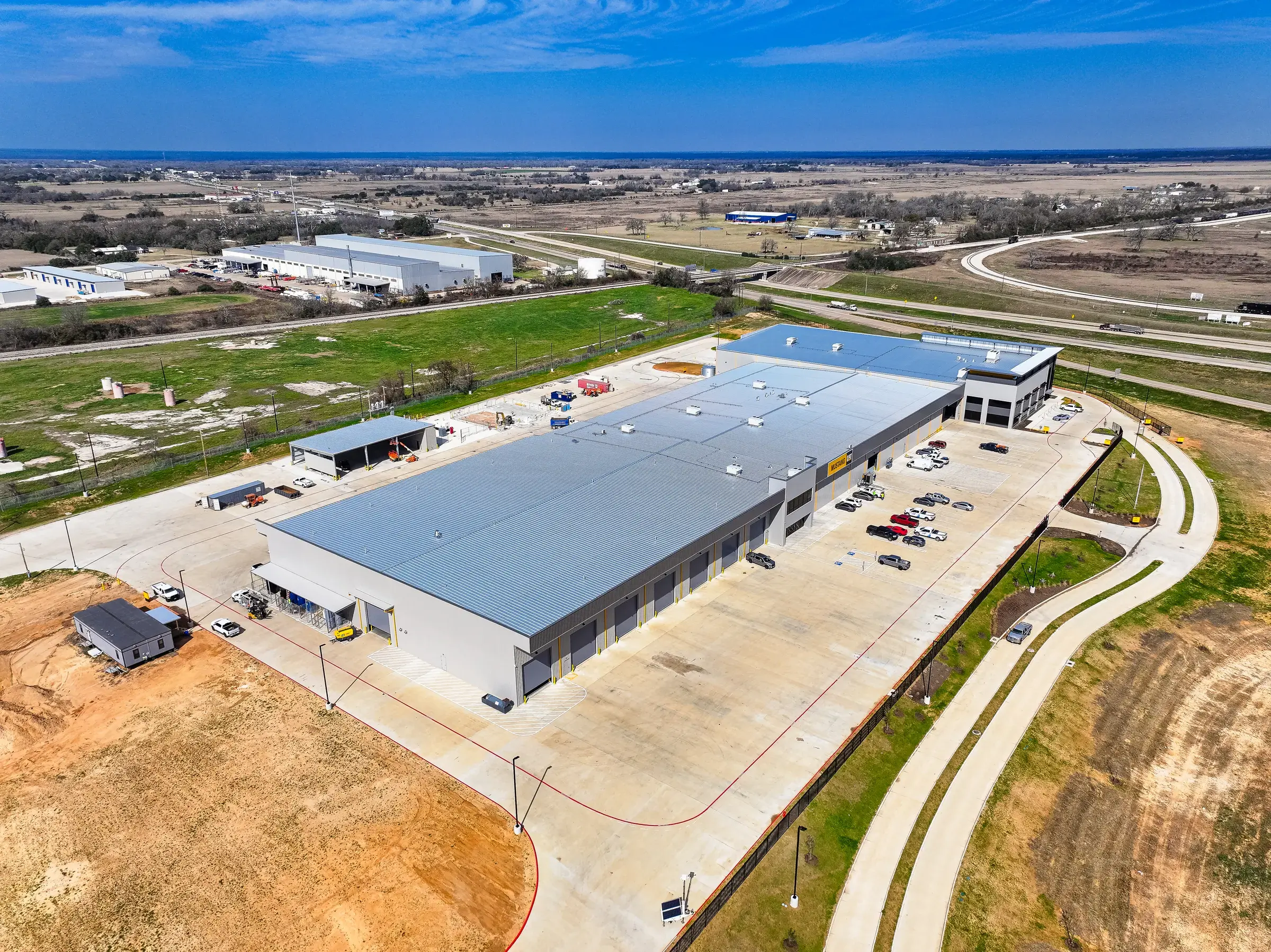 Aerial view of the completed facility and its concrete yard, set in open farmland with the highway beyond