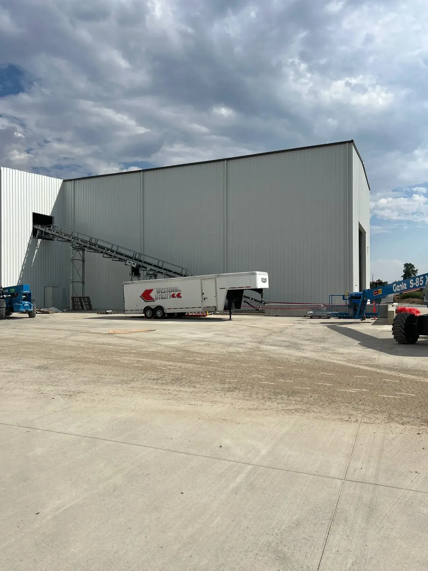 The conveyor running from the batch plant into the building at the paved apron, with material handling equipment staged alongside
