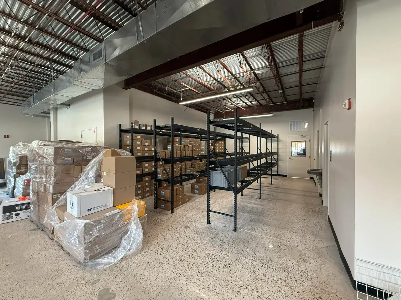 The warehouse side of the building — pallet racking and stock along one wall beneath exposed structure, new ductwork, and linear light fixtures